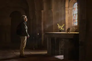Elderly woman stands quietly in a dim Romanesque chapel at dawn, illuminated by soft colored light from stained glass onto volcanic stone near the altar.