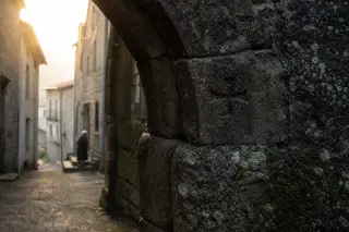 Medieval basalt archway in Pradelles at dawn, dew on the stones and a faint cross carving, elderly woman near her doorway in soft misty light.