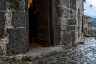 Basalt stone threshold and partially open wooden door of an old Pradelles house with an elderly silhouette inside, rainy cobblestone street outside, soft cloudy afternoon light.
