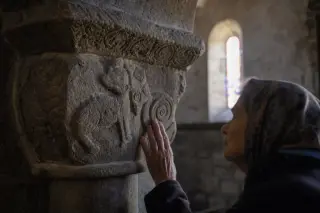 Romanesque stone capital with sculpted details, touched by soft morning light, elderly woman in background contemplating