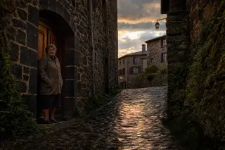 Elderly woman in a wool cardigan standing on a stone doorstep along a narrow, glistening cobbled alley of Pradelles at sunset, with golden light and wet basalt walls.