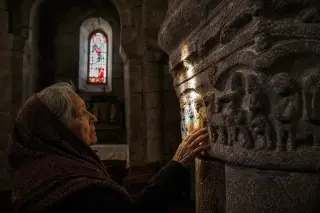Rain-darkened basalt stone steps in a medieval Pradelles alley with an elderly woman in a shawl standing in a stone doorway, under soft, gray daylight