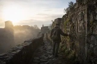 Solitary hiker with worn backpack pauses at basalt rampart in Pradelles, dawn light skimming ancient stone textures and morning mist lingering along the walkway.