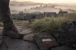 Low-angle view of a hiker’s worn boot and trouser edge beside a basalt path with dew and a faded GR marker, misty rooftops and ancient walls of Pradelles in the background, capturing a quiet dawn before setting out.