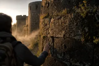 Weathered volcanic stone wall of medieval fortifications at dawn, touched by golden light and light mist, with a solitary hiker pausing in contemplation, partially visible in the frame.