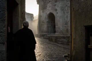 Mist-filled medieval alley in Pradelles at dawn, elderly local woman in silhouette pausing at her stone doorway, soft golden morning light highlighting carved symbols above an ancient porch.